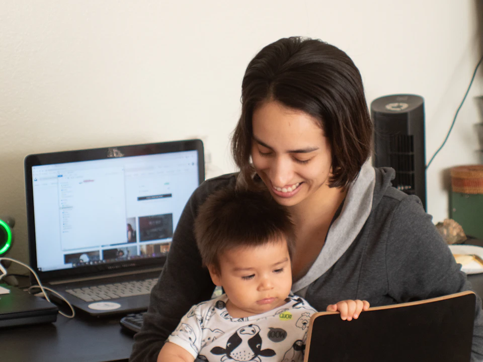 Eine Mutter sitzt mit ihrem Baby auf dem Schoß lächelnd vor einem Laptop.